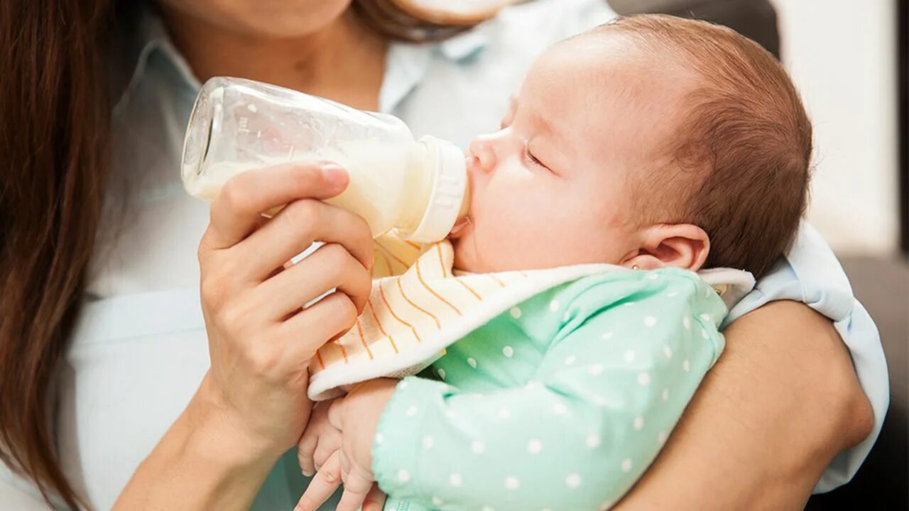 Мама с младенцем держит в руках бутылочку. Младенец срыгивает много молока утром. Новорожденный икает. Новорожденный часто срыгивает после кормления. Часто срыгивает после