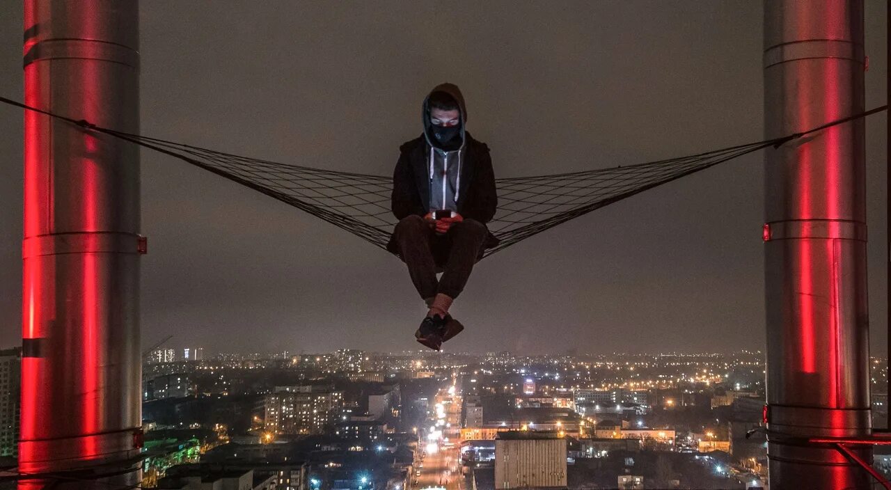 Человек стоит над городом. Человек на небоскребе. Руферы на крыше. Человек сидит на крыше.