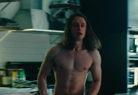 A shirtless and muscled Rory Culkin stands in the middle of a grey kitchen....