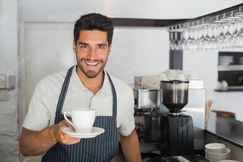 Парень с кофе улыбается. Парень на кухне улыбается. Мужчина протягивает кофе. Бариста улыбается.