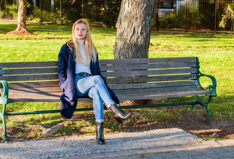 Девушка с телефоном в парке днем. Блондинка сидит на скамейке задом. A woman is sitting on a Bench and smoking. Sit on a bench