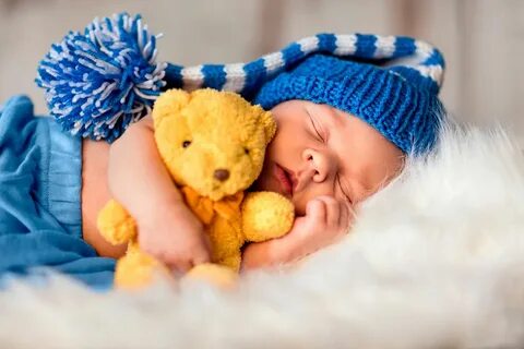 a baby sleeping next to a teddy bear wearing a blue hat and holding a yello...