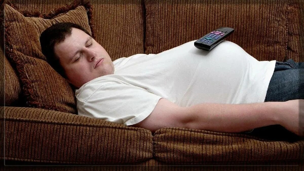 Толстый человек на диване. Человек лежит на диване. Малоподвижный человек. Низкая физическая активность.
