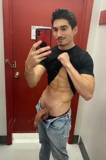 Handsome Latino guy standing in a dressing room, pulling up his shirt while...