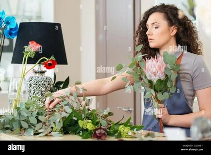 Cheerful lovely young woman florist choosing flowers and making bouquet in shop...