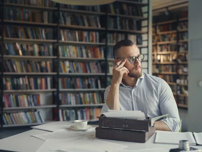 Мужчина сидит за столом смотрит на бумаги. Писатель-профессионал. Человек за работой. Обложка для веб студии. Обложка для статьи.