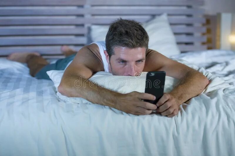 Лежа играть в телефон. Человек лежит на кровати с телефоном. Мужчина лежит на кровати. Парень с телефоном в кровати. Телефон лежит.