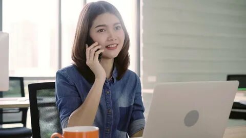 A beautiful young smiling Asian woman is working on a laptop when she answe...